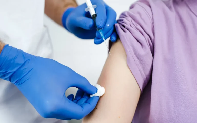 Zorgverlener met blauwe handschoenen geeft een vaccinatie in de bovenarm van een vrouw in een lila shirt