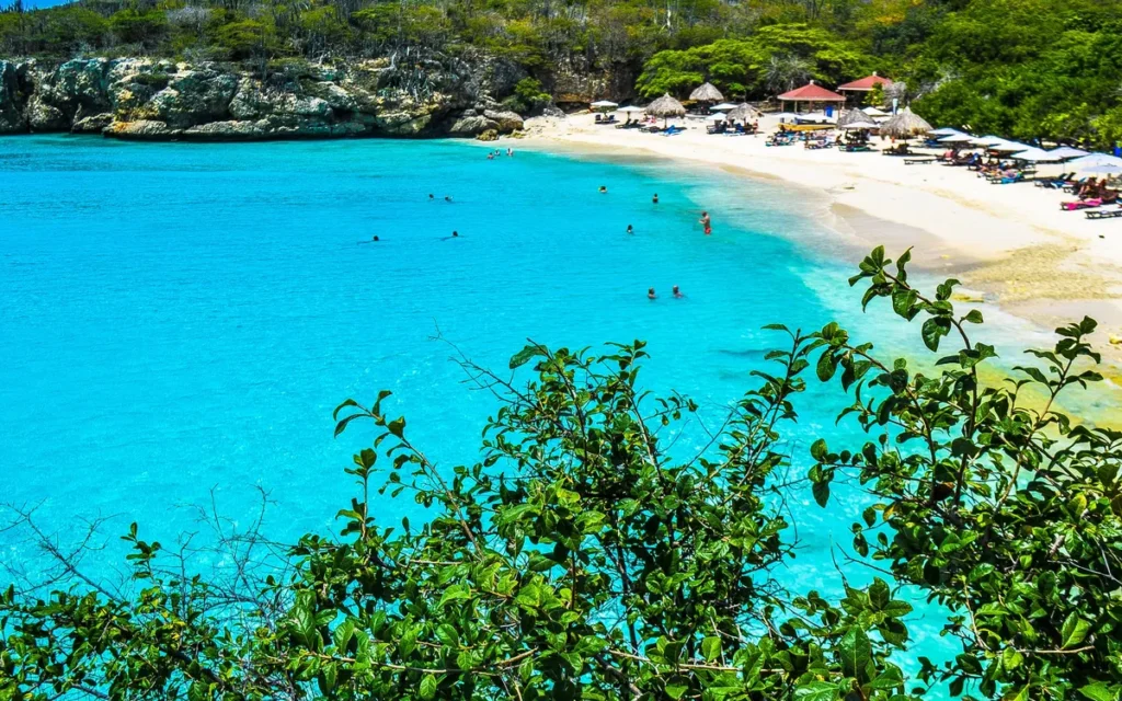 Baai met wit zandstrand, turquoise water en strandparasols tussen groene kliffen op Curaçao