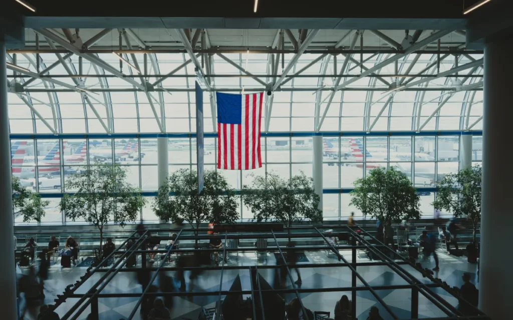 Vertrekhal van een Amerikaans vliegveld met een grote Amerikaanse vlag voor de glazen gevel en vliegtuigen buiten